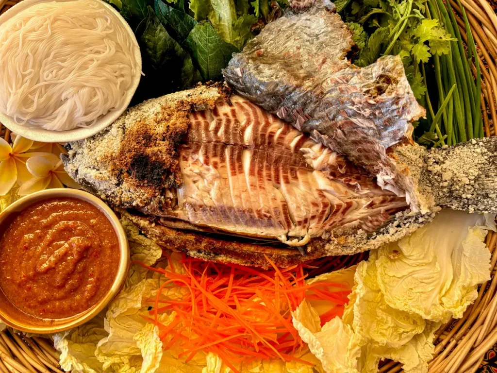 Pla pao, authentic Thai grilled fish, served with a seafood dipping sauce, fresh vegetables, and rice noodles.