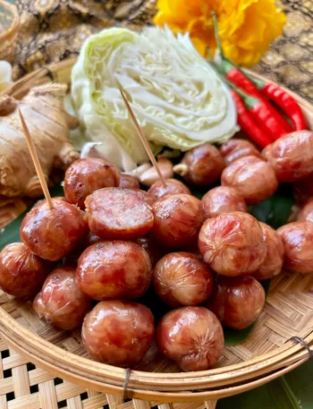 Sai krok Isan Thai sausages on a bamboo basket, with a backdrop of chilies, ginger, and cabbage.