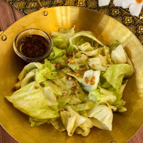 Golden wok filled with Thai cabbage stir-fry, garnished with fried garlic, served with chili oil on the side.