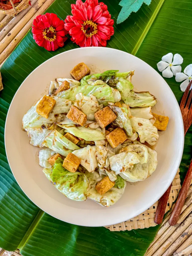 Overhead shot of Thai-style fried cabbage with golden tofu, styled with flowers and banana leaf backdrop.