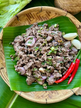 Thai ground beef larb with shallots, fresh herbs, and chilies resting on a banana leaf set on a bamboo plate, with garlic cloves to the side.