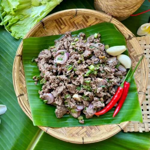 Thai ground beef larb with shallots, fresh herbs, and chilies resting on a banana leaf set on a bamboo plate, with garlic cloves to the side.