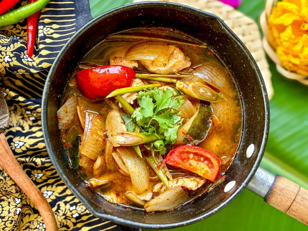 Thai lemongrass soup with chicken, tomato, onion, kaffir lime leaves, and herbs in a black pot.