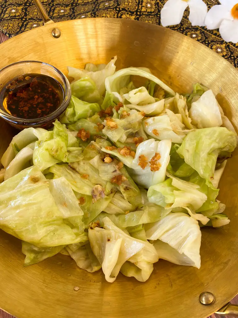Thai sautéed cabbage in a wok, ready to be served, featuring golden fried garlic and a side of spicy chili oil.