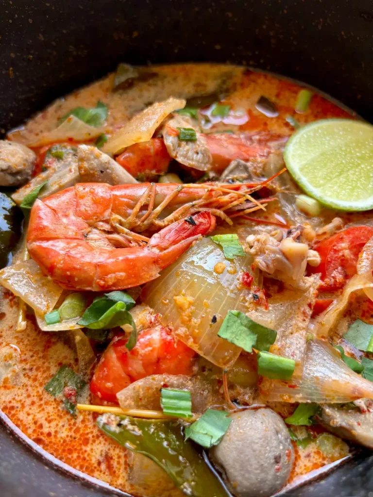 Close-up of Thai seafood soup showcasing prawns, mushrooms, tomatoes, onion, lemongrass, galangal, garnished with a lime slice.