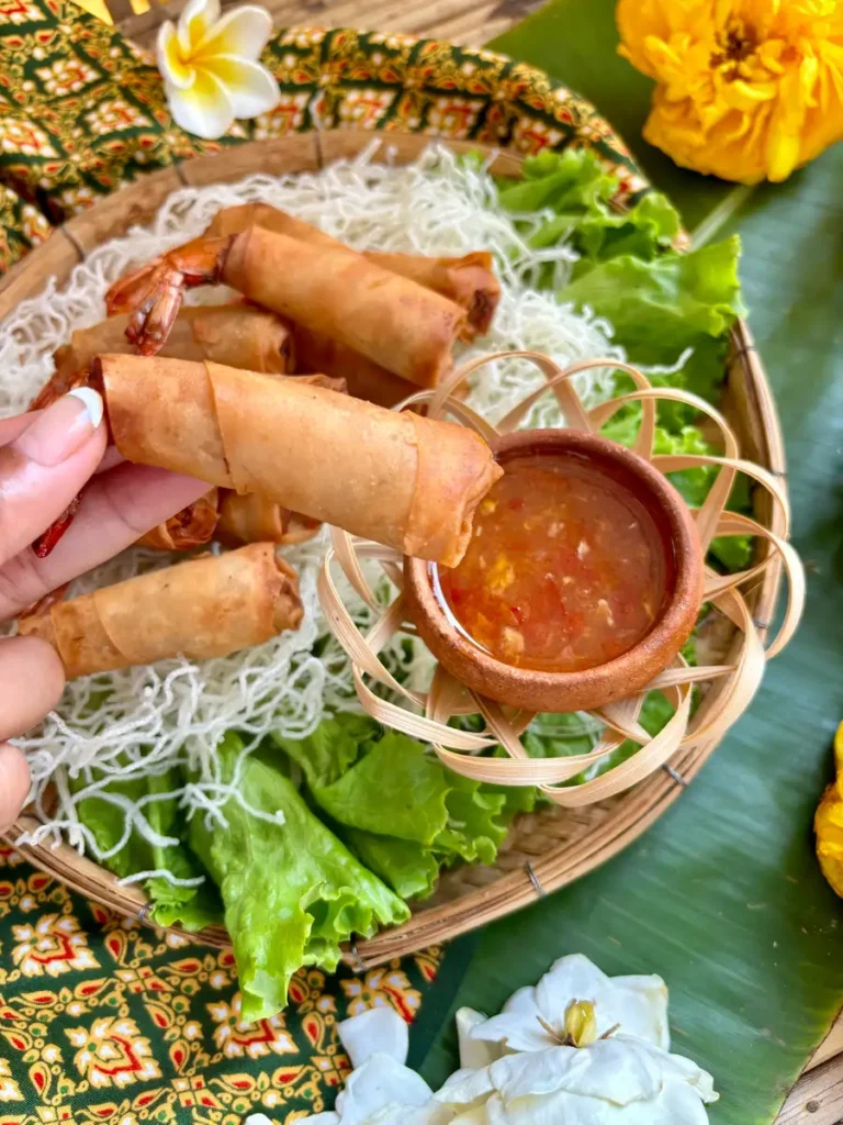 Hand picking up a crispy Thai shrimp roll with fresh herbs and dipping sauce backdrop.