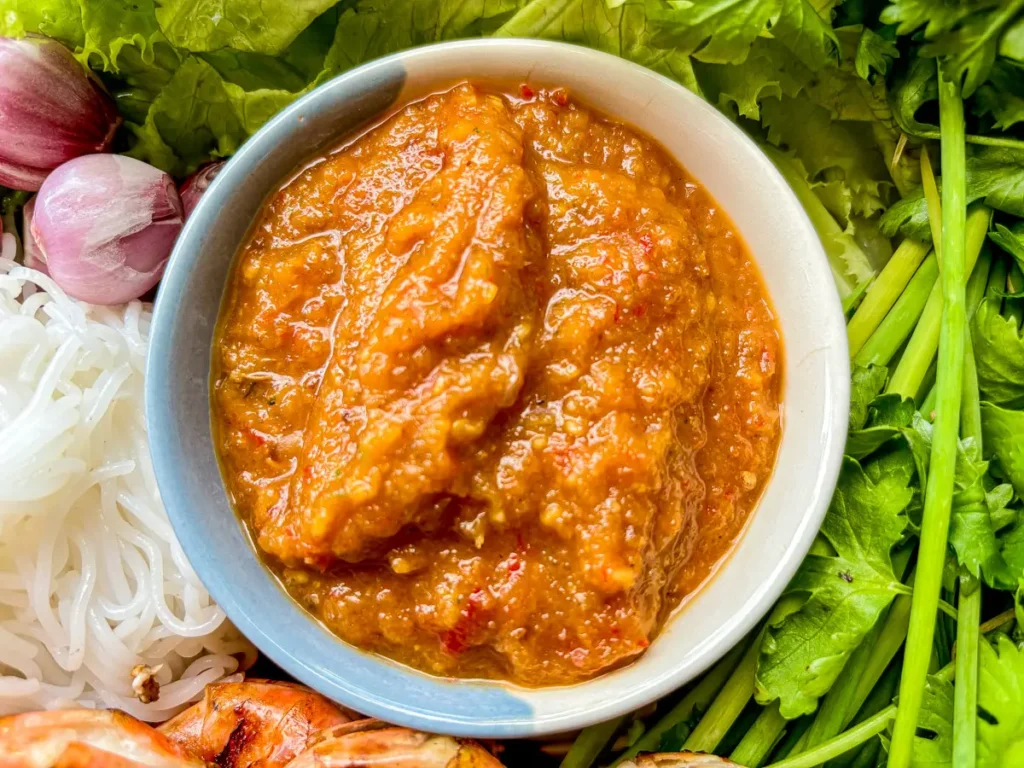 Close-up of Thai nam jim tua spicy peanut dipping sauce with a rich orange color, served with herbs, noodles, and grilled prawns.