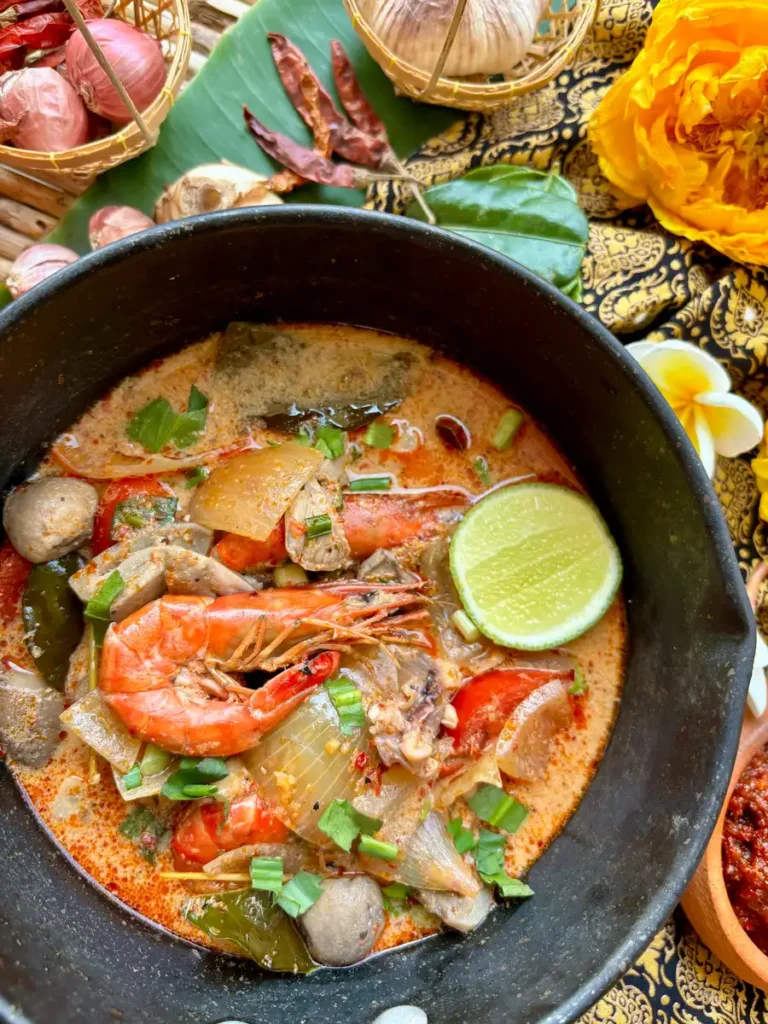 Authentic Thai tom yum talay soup in a rustic pot, adorned with a lime wedge and surrounded by traditional ingredients and fresh herbs and spices.