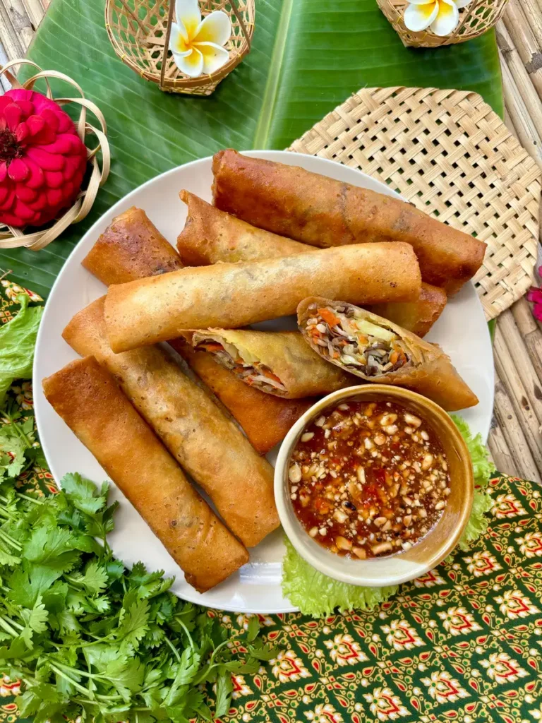 Plate of golden-brown fried Thai vegetable spring rolls with fresh herbs and chili dipping sauce.