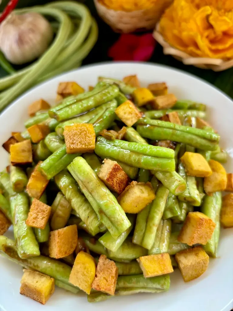 Tofu and long bean stir-fry served in a white dish, with a backdrop of more Chinese long beans.