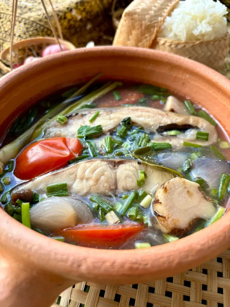 A traditional clay pot filled with tom pla, a Thai fish soup, showcasing slices of fish, tomatoes, and chopped green onions, served next to a basket of sticky rice.