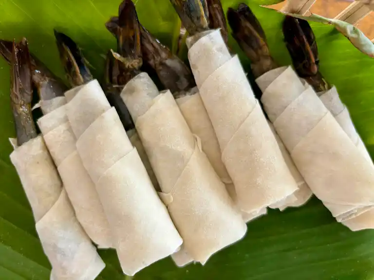 Uncooked shrimp in a blanket wrapped in spring roll wrappers on a banana leaf.