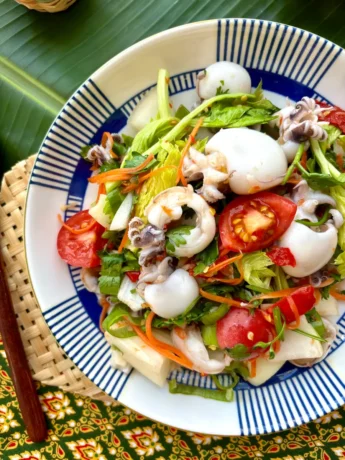 Yum pla muk, a Thai squid salad with vegetables, served on a porcelain plate, arranged on a banana leaf.