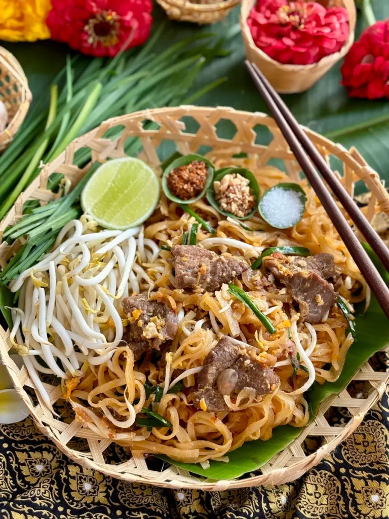 Authentic beef pad Thai on a bamboo dish with lime, garlic chives, chili flakes, and crushed peanuts.