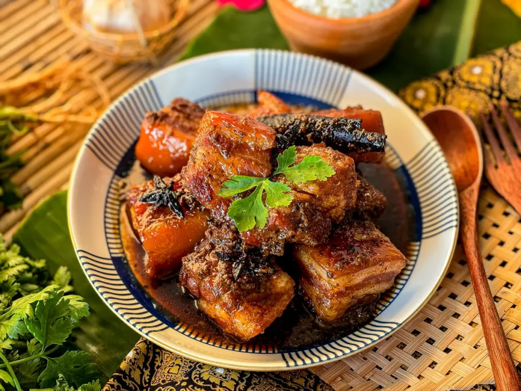 Moo hong, Thai pork belly stew, with spices served in a deep bowl, surrounded by herbs and a bowl of jasmine rice.