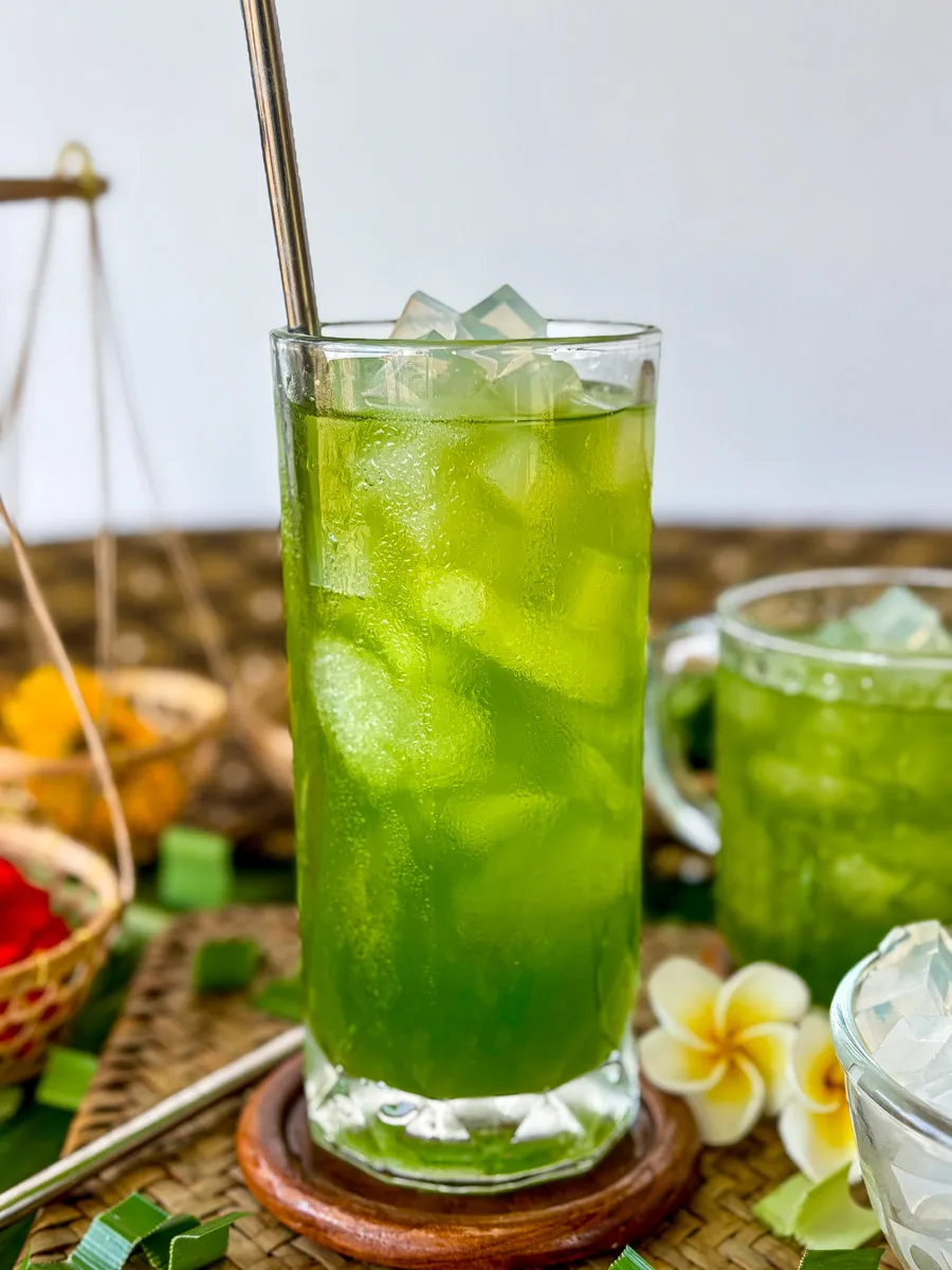 Thai pandan drink served in a tall glass with ice and metal straw, featuring bright green color and chilled cubes of jelly.
