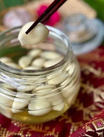 Pickled garlic in glass jar with clear brine and chopsticks lifting a single clove.