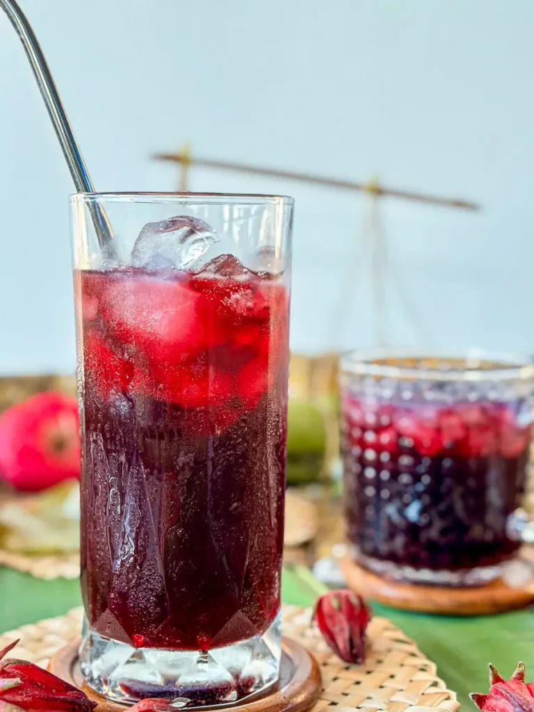 Tall glass of chilled roselle drink with ice cubes and metal straw, served on a rustic Thai-style setup.