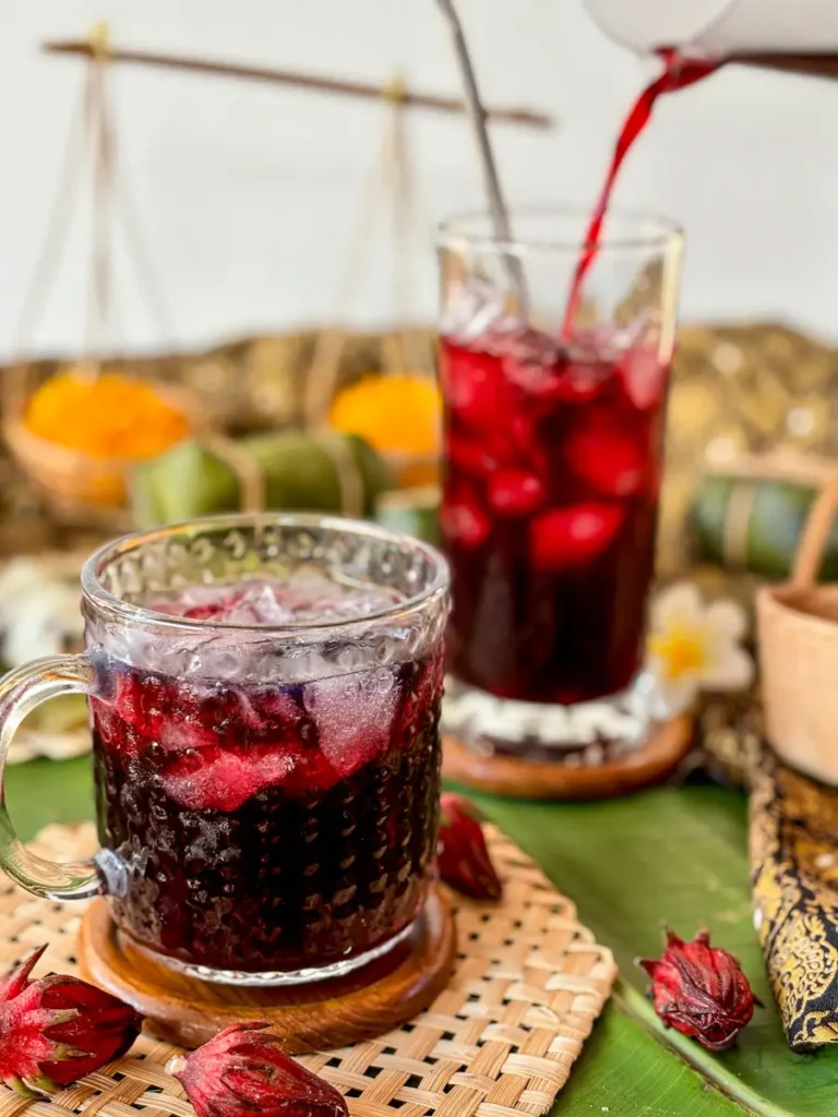 Pouring vibrant red roselle juice into a glass over ice, surrounded by dried roselle buds and Thai décor.