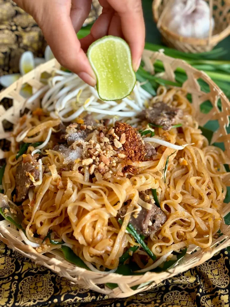 Hand squeezing lime over beef pad Thai garnished with bean sprouts and peanuts.