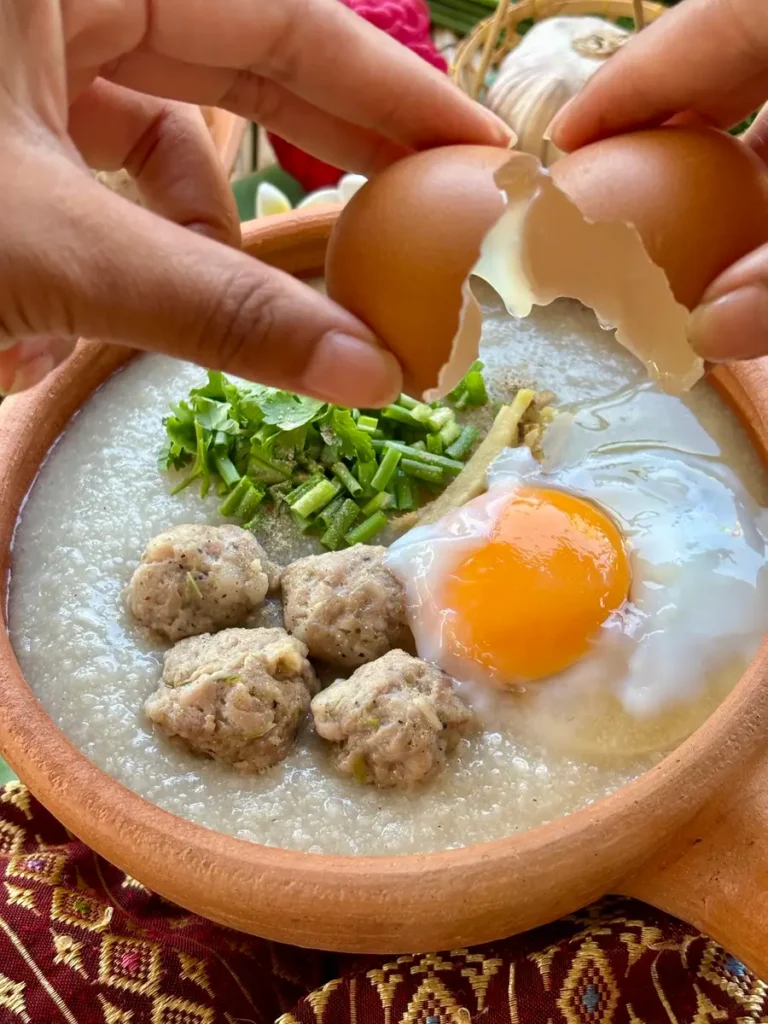 Adding an egg to a steaming bowl of Thai jok for classic rice porridge with pork meatballs.