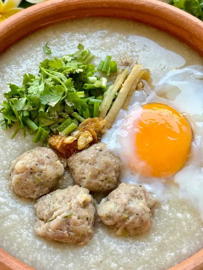Close-up of Thai rice porridge, jok, garnished with fresh herbs, pork meatballs, and an egg in a clay pot.