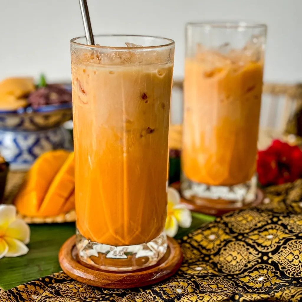 Glass of iced Thai tea, cha yen, with milk and ice, served with a metal straw on a traditional patterned cloth.