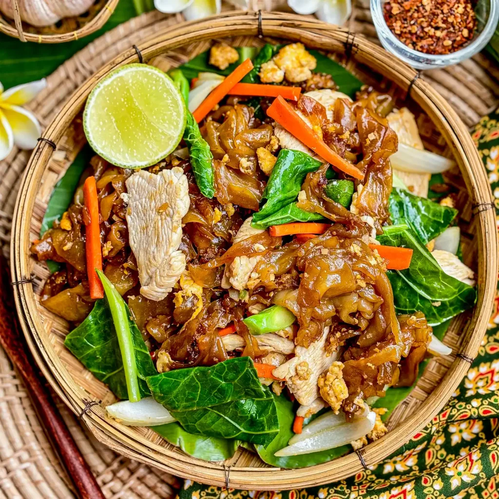 Thai chicken pad see ew served in a traditional bamboo basket with lime wedge and colorful vegetables.