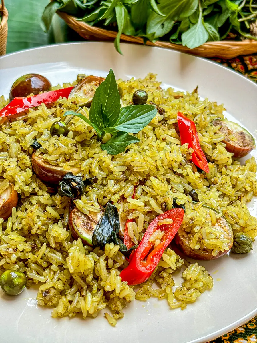 Close-up of green curry fried rice with fresh herbs, sliced red chili, and Thai eggplants on a patterned fabric.