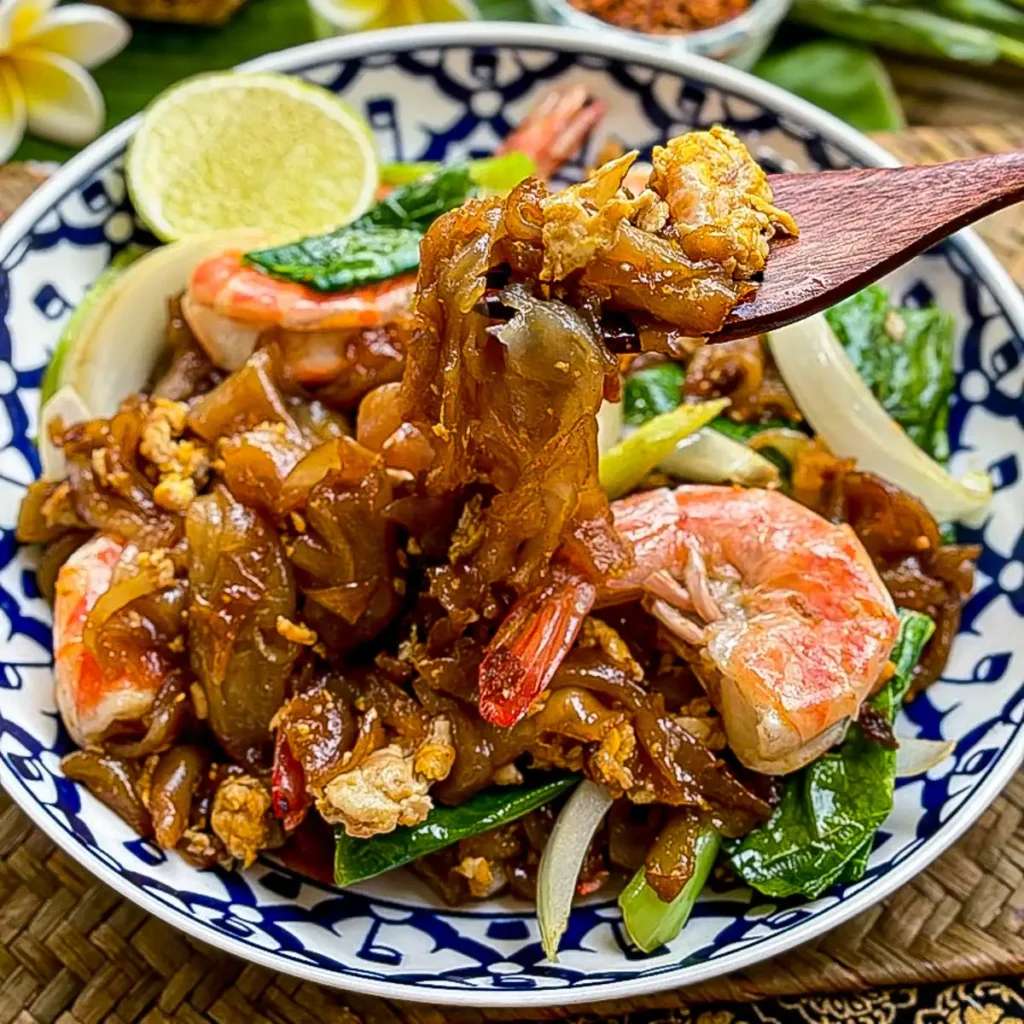 Spoon lifting chewy rice noodles from a bowl of shrimp pad see ew, with egg, greens, and onions in a dark soy sauce glaze.