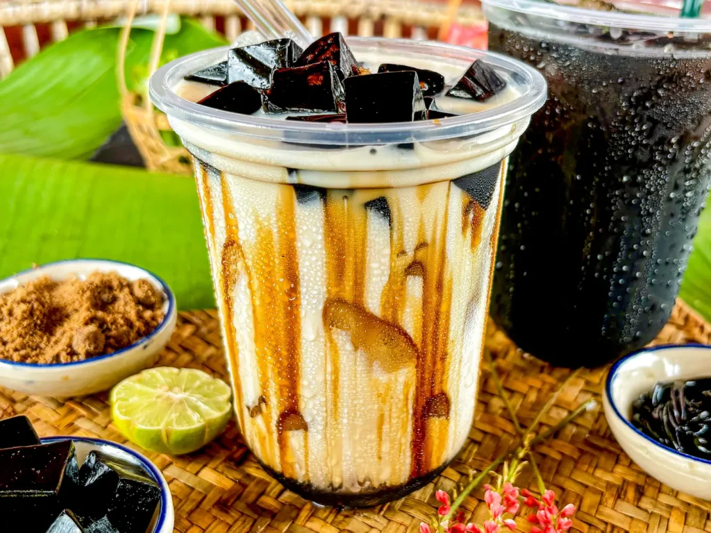 Grass jelly boba with brown sugar syrup served in a clear plastic cup, topped with glossy grass jelly cubes and surrounded by lime, brown sugar, and syrup.