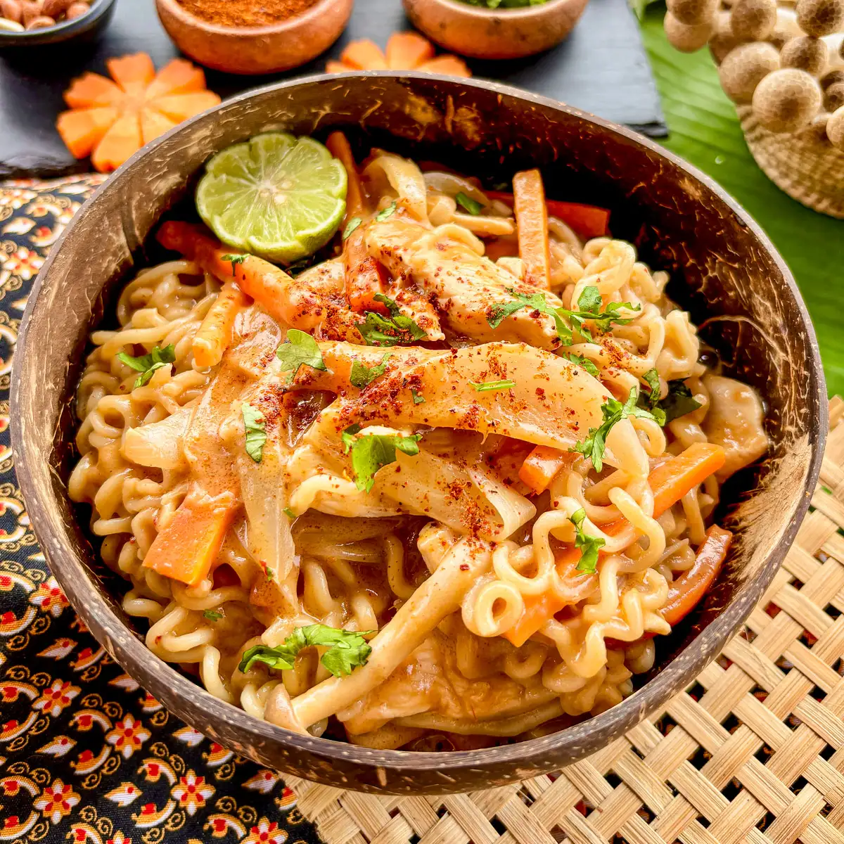 Bowl of Thai peanut butter ramen noodles with chicken, vegetables, and fresh herbs, served with lime and chili flakes.