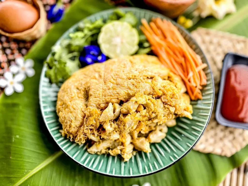 Thai crab omelette served on a green plate with fresh herbs, lime, sliced carrots, and chili sauce.