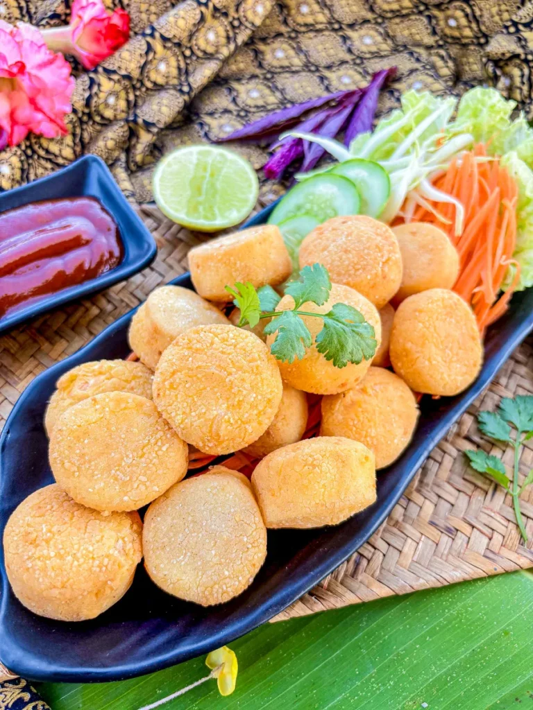 Crispy fried egg tofu pieces served with shredded vegetables, cucumber, and dipping sauce on a black plate over a woven mat and banana leaf.