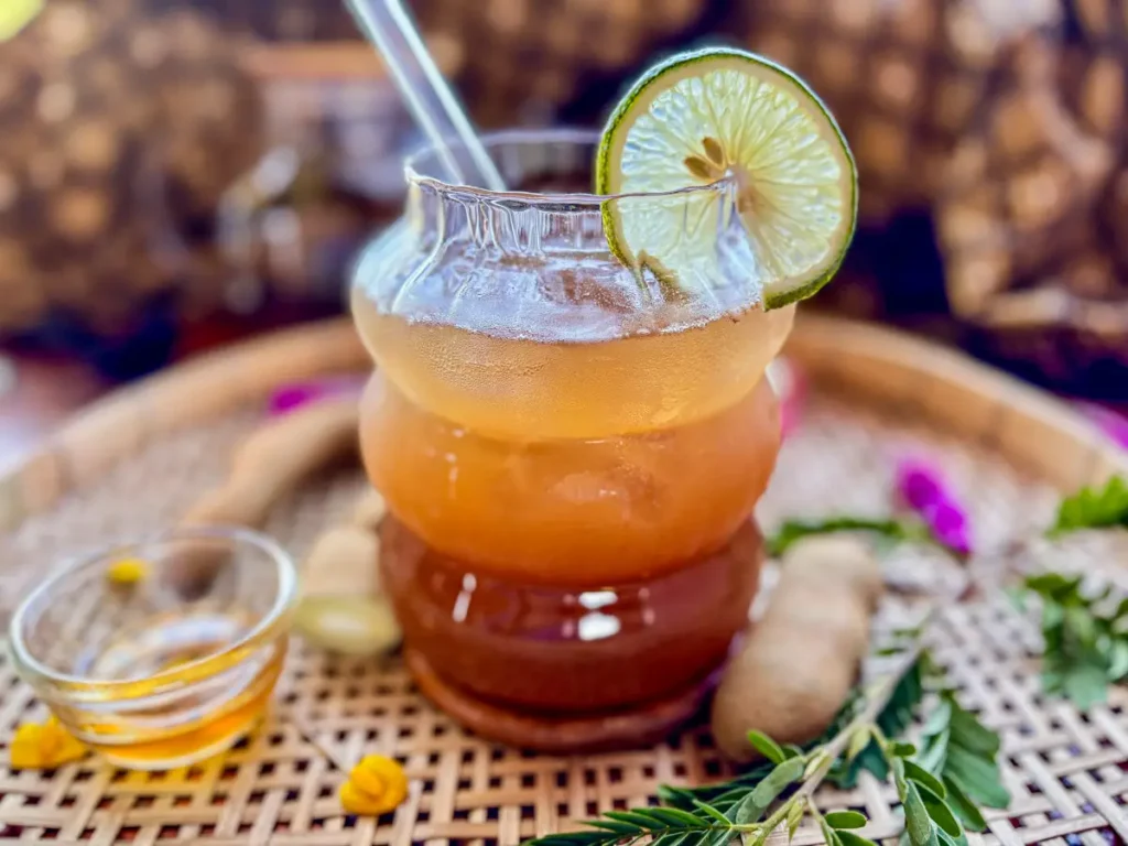Glass of tamarind iced tea with a slice of lime, served on a woven tray with honey and fresh tamarind pods.
