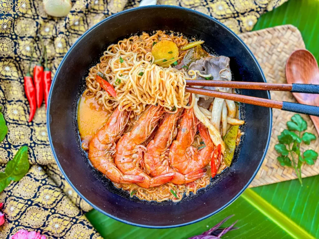 A close-up of tom yum mama noodles with shrimp, garnished with fresh herbs and served in a black bowl with a rich, flavorful broth.