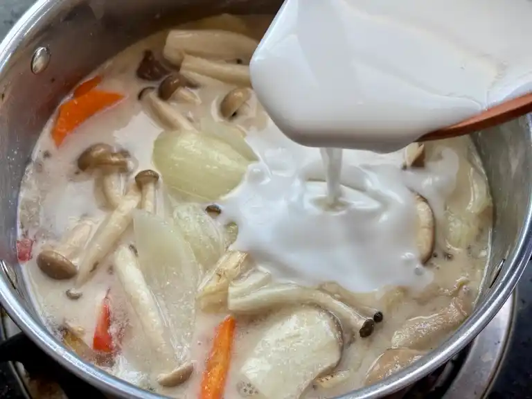 Pour coconut milk into a pot of tom kha hed with mushrooms and vegetables.