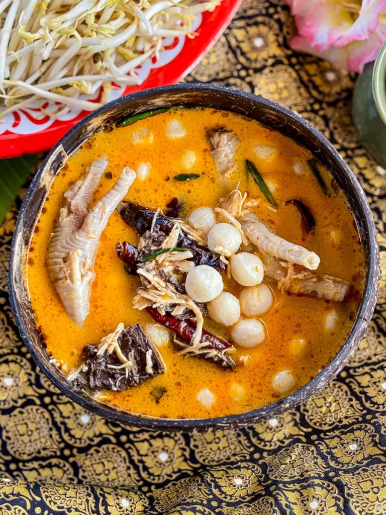 A close-up of Thai chicken feet curry in a coconut shell bowl, highlighting the creamy red curry broth and fresh vegetables.