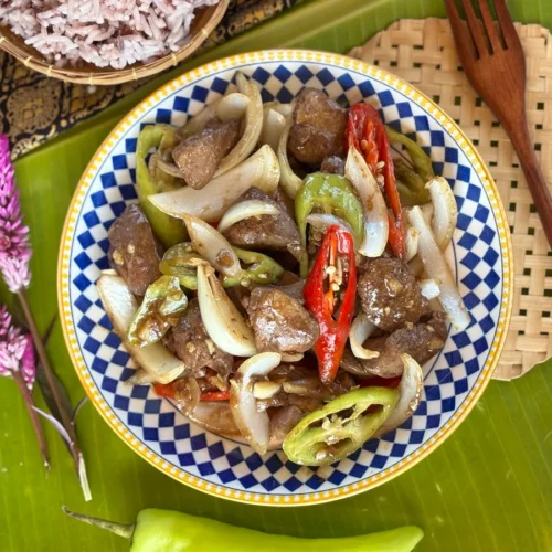 Overhead photo of stir-fried chicken livers garnished with chili peppers, served with rice on a placemat with banana leaves.