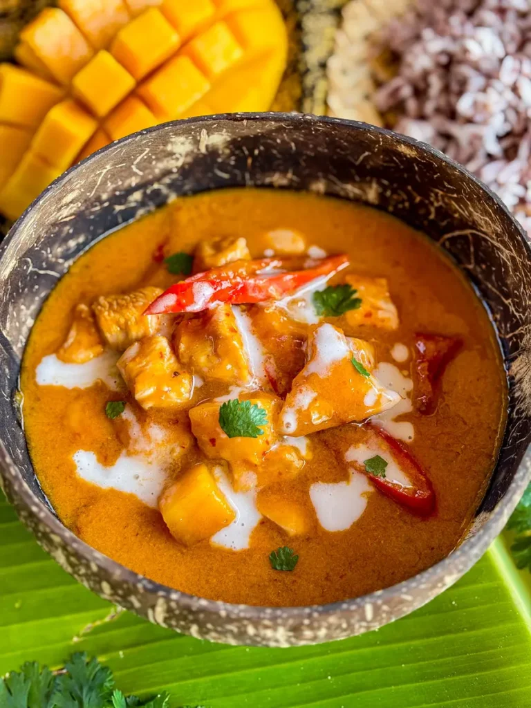 Thai mango chicken curry in a coconut bowl garnished with fresh cilantro and red chili, with a fresh mango in the background.
