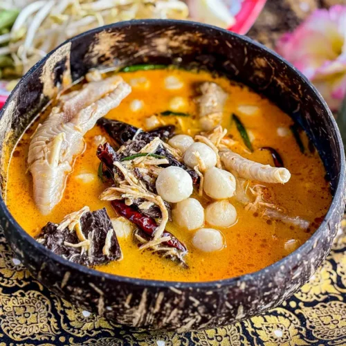 Thai chicken feet curry served in a coconut shell bowl, with a rich red curry sauce and garnished with fish balls and vegetables.