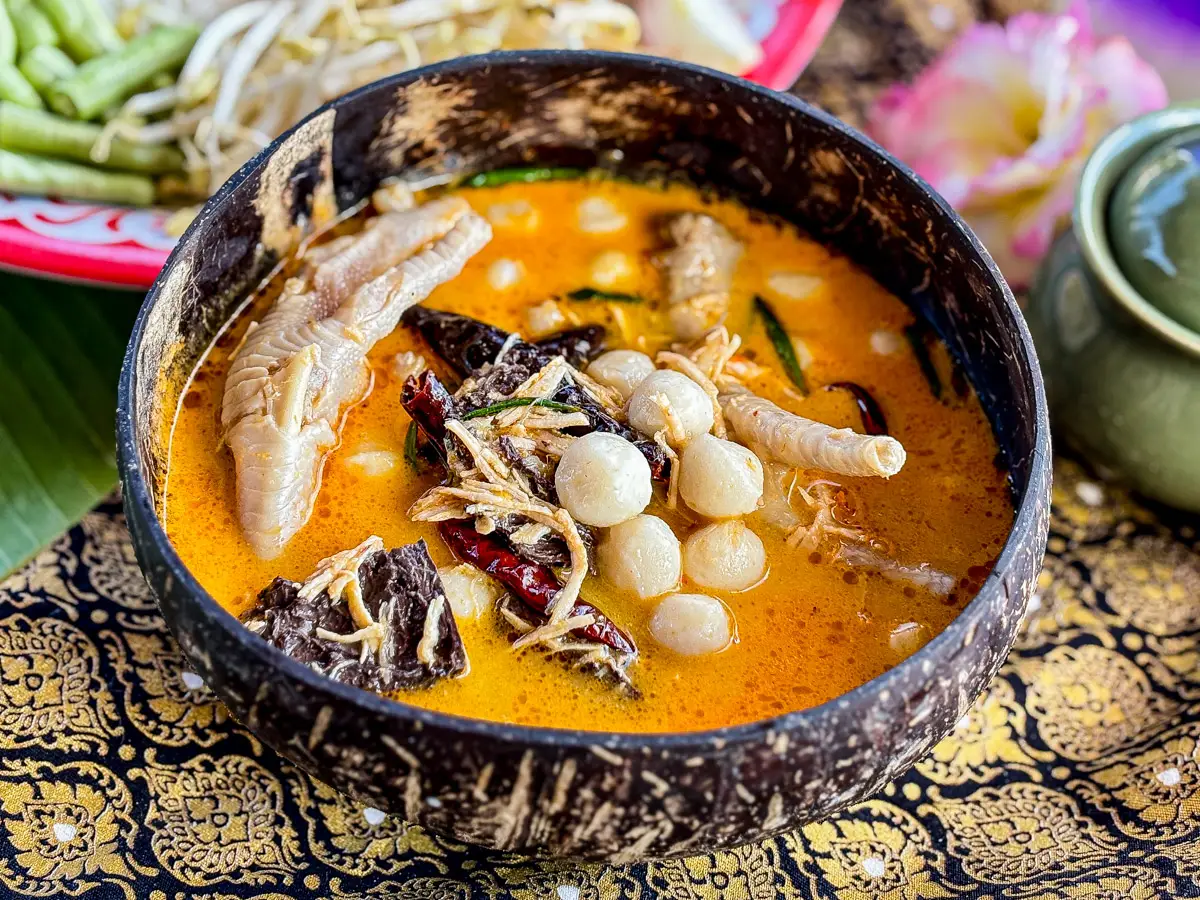Thai chicken feet curry served in a coconut shell bowl, with a rich red curry sauce and garnished with fish balls and vegetables.