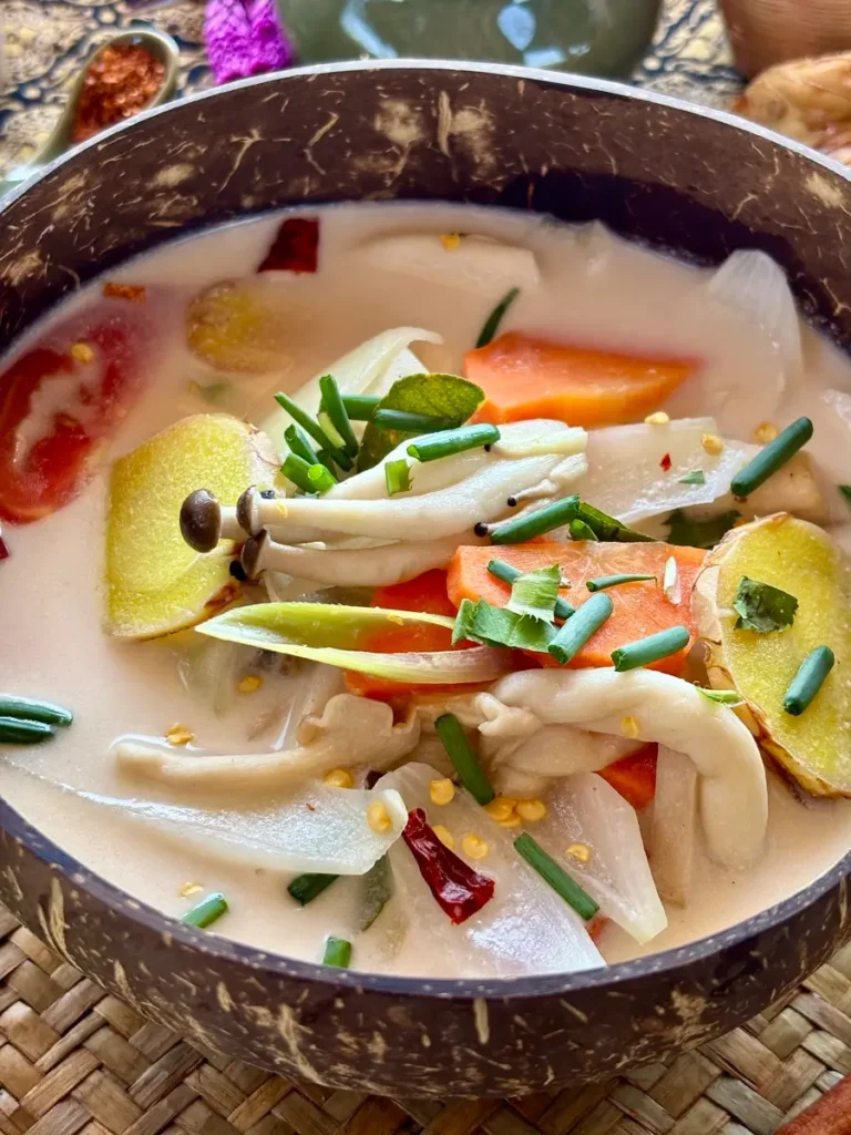 Tom kha hed, a creamy Thai mushroom vegetarian soup in a coconut shell bowl, garnished with green onions and chilli flakes.