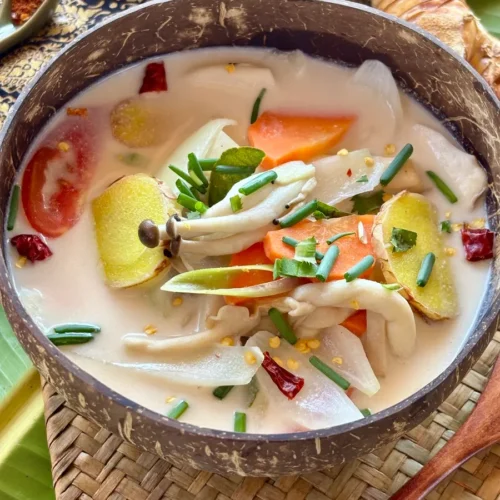 A bowl of creamy vegan Thai mushroom soup with coconut milk and vegetables, served in a coconut shell on a woven mat with garnishes.