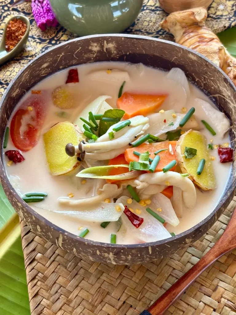 A bowl of creamy vegan Thai mushroom soup with coconut milk and vegetables, served in a coconut shell on a woven mat with garnishes.