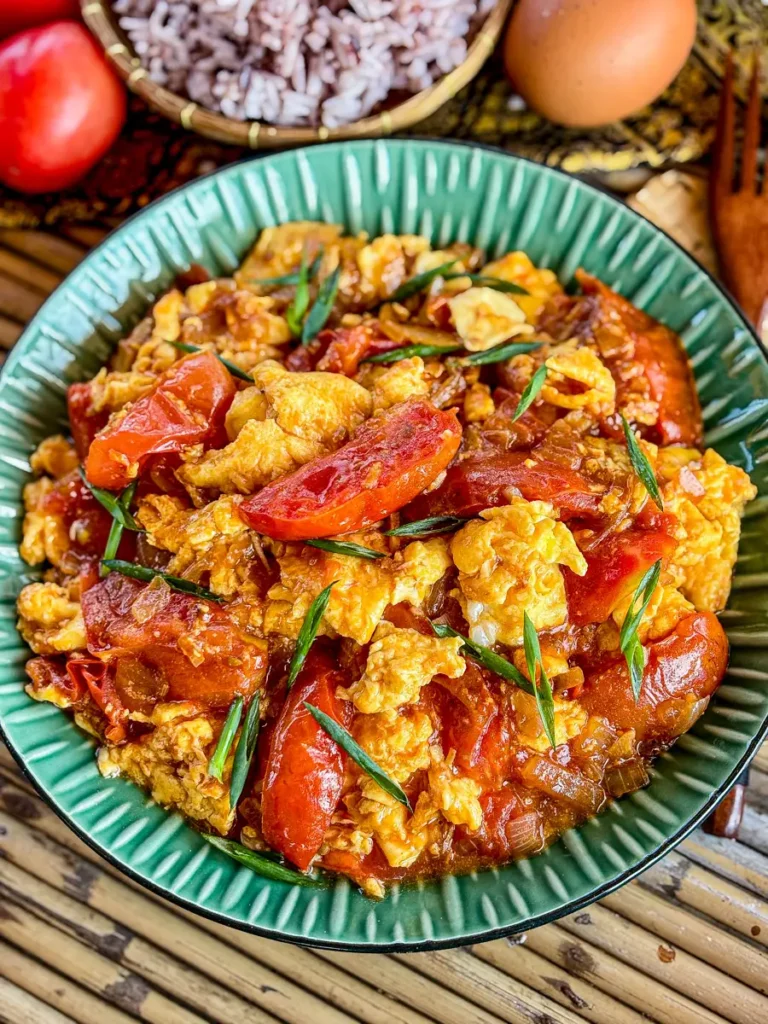 A bowl of Thai tomato and egg stir-fry garnished with green onions, served alongside a bowl of rice, fresh tomatoes, and eggs.