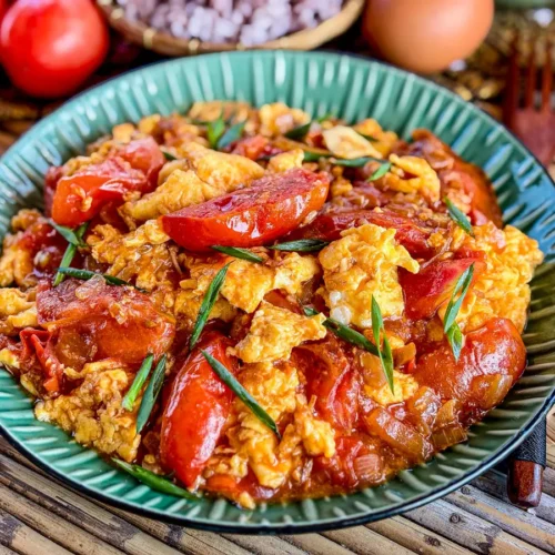 Thai-style tomato and egg stir-fry in a green plate, showcasing vibrant red tomatoes, fluffy scrambled eggs, and green onion garnish.