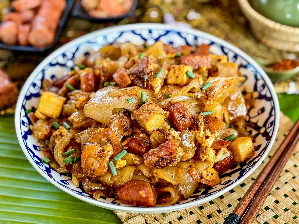 A bowl of cry baby noodles with a mix of tofu, sausage, egg, and rice noodles, placed on a banana leaf with chopsticks.