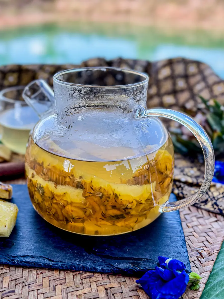 A glass teapot filled with pineapple skin tea, surrounded by pineapple chunks, cinnamon, and butterfly pea flowers, with a serene outdoor backdrop.