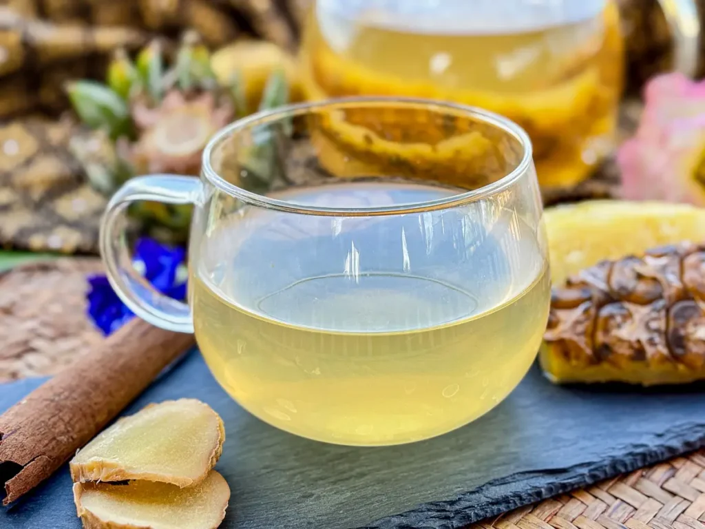 Pineapple tea served with fresh ginger slices, a cinnamon stick, and a teapot filled with pineapple skin and core in the background.